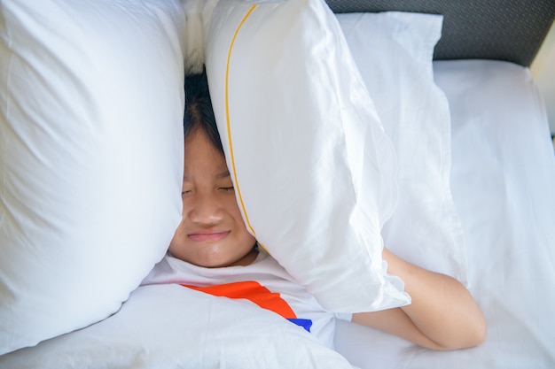 Bambina sdraiata a letto che copre la testa con il cuscino perché rumore fastidioso troppo forte. bambino irritato che soffre di vicini rumorosi, che cerca di dormire dopo il segnale di sveglia dell'allarme