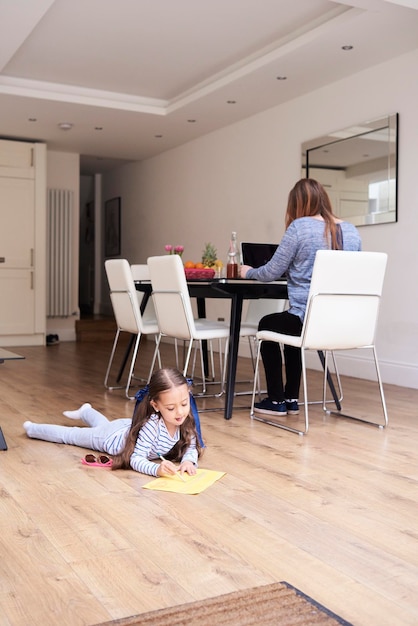 Little girl lying on floor drawing a picture while her mother working on laptop in the background