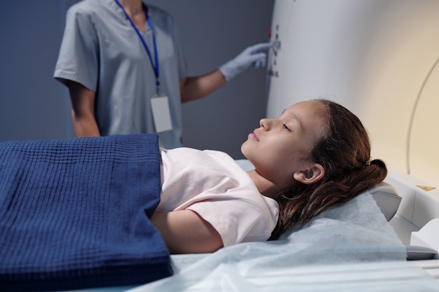 Little girl lying on table of mri scan machine while gloved assistant pressing button on panel