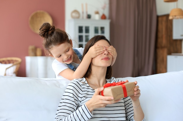 Little girl make birthday present to excited parent , smiling little child congratulate give gift box to happy mother and closed mother's eyes.