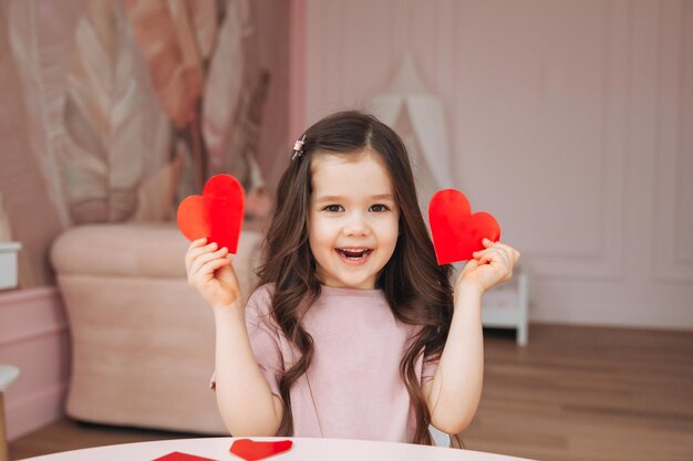 A little girl makes Valentine's Day cards using colored paper scissors and pencil sitting at a table