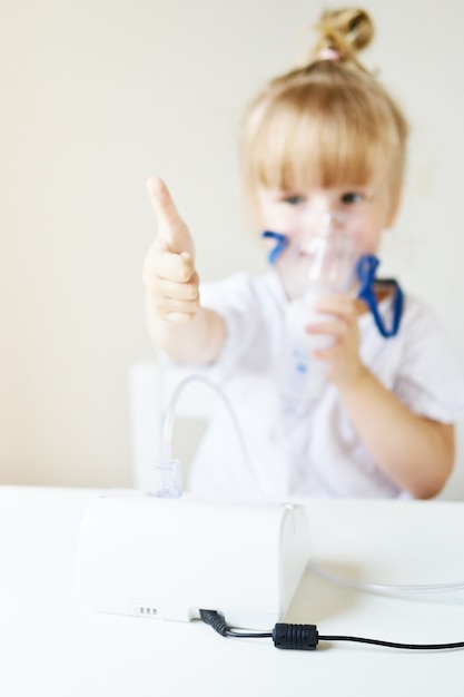 Little girl in a mask for inhalations, making inhalation with nebulizer at home inhaler on the table, indoor, sick child