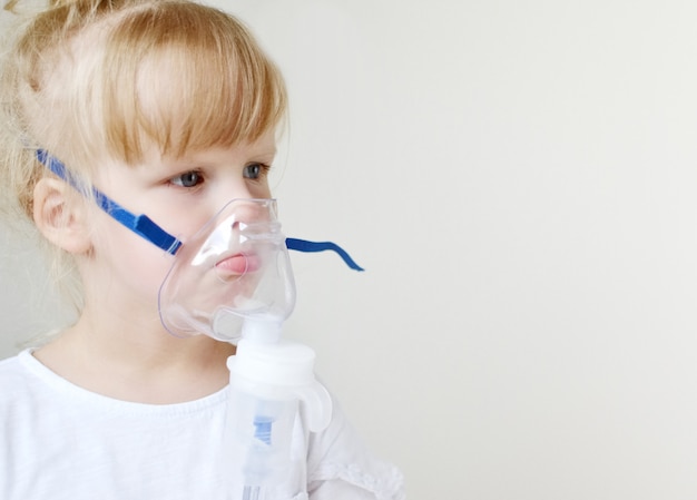 Little girl in a mask for inhalations, making inhalation with nebulizer at home inhaler on the table, indoor, sick child