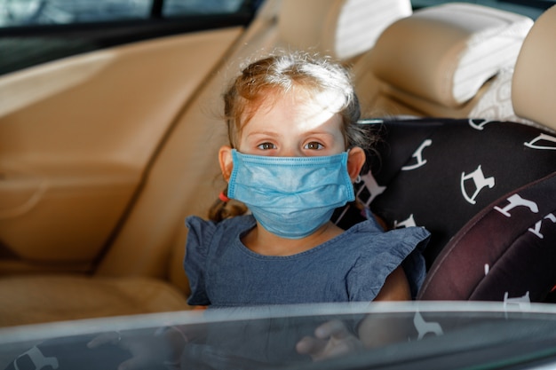 Little girl in a medical mask is sitting in a child seat in the car.