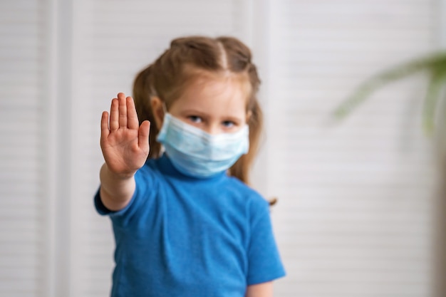 Little girl in a medical mask looks at camera and makes a stop gesture