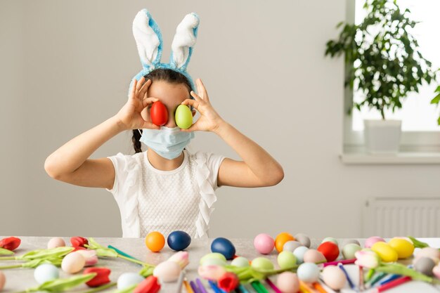 Little girl in a medical mask paints Easter eggs with a pattern. Stay home.