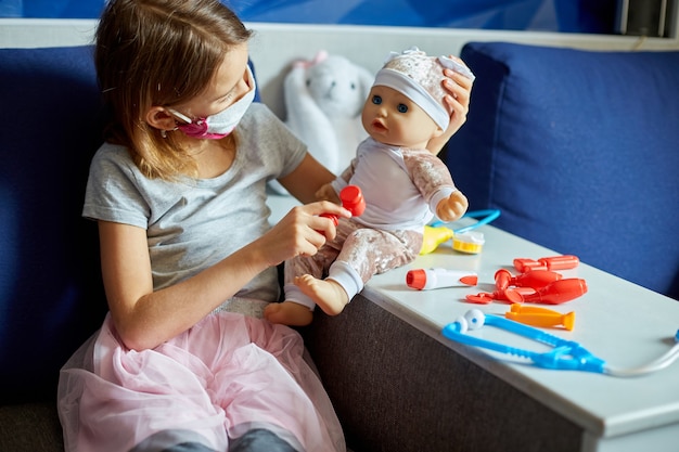 Una bambina in maschera medica si siede sul divano, interpreta un dottore, cura e ascolta la bambola con uno stetoscopio in maschera medica, a casa durante la quarantena e la pandemia