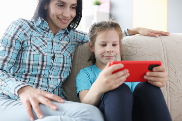 Little girl and mother are sitting on couch with mobile phone in their hands