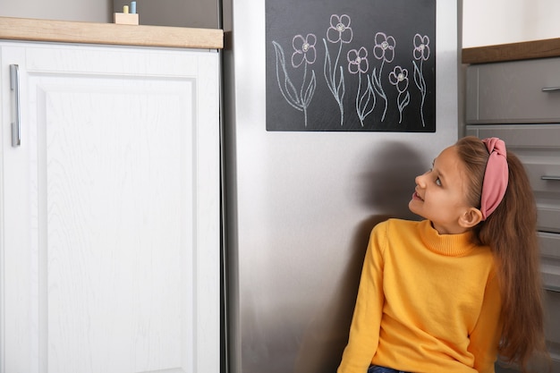 Little girl near chalkboard on refrigerator in kitchen