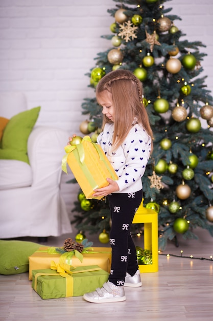 Маленькая девочка возле елки открывает рождественские подарки