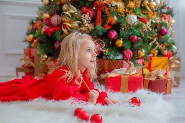 A little girl near the Christmas tree writes a letter to Santa Claus at home