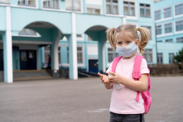 A little girl near the school with a backpack in a mask and an antiseptic in her hands the concept