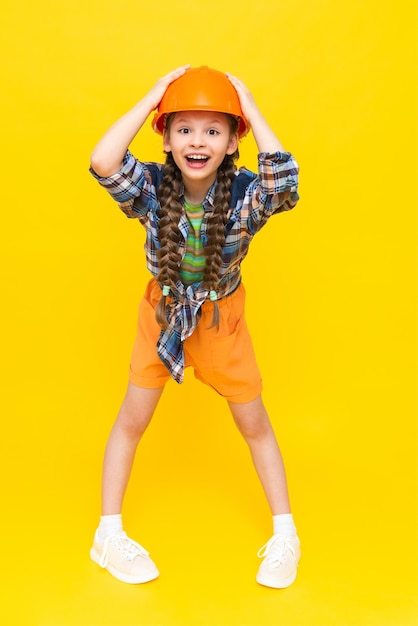 A little girl in an orange construction helmet is holding her head and smiling broadly happily The child is preparing for repairs in the nursery In a mouthful Vertical Yellow isolated background
