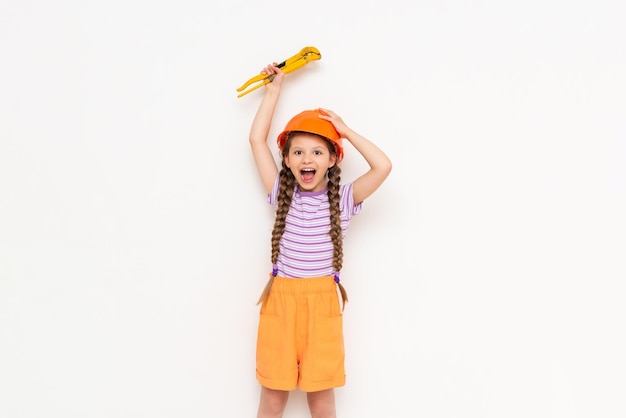 A little girl in an orange construction helmet with two pigtails raises a monkey wrench and smiles broadly on a white isolated background The concept of renovation in the children's room