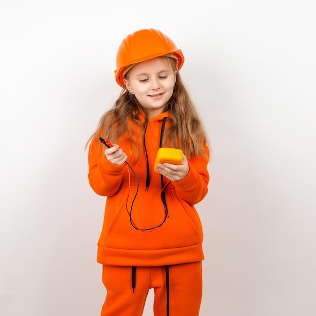 Una bambina in una tuta arancione e un casco da costruzione in possesso di un concetto multimetro di un giovane elettricista sfondo bianco ritratto festa del lavoro