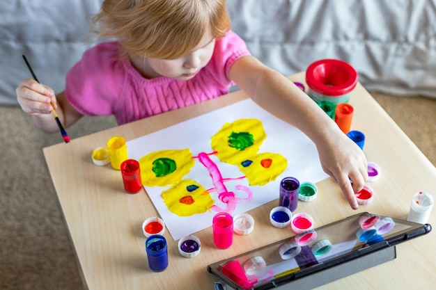 Фото Маленькая девочка рисует с красками, глядя на планшет, сидя за столом в домашней онлайн-школе