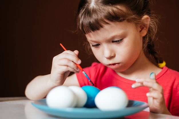 Little girl paints Easter eggs on blue dish sitting at the table