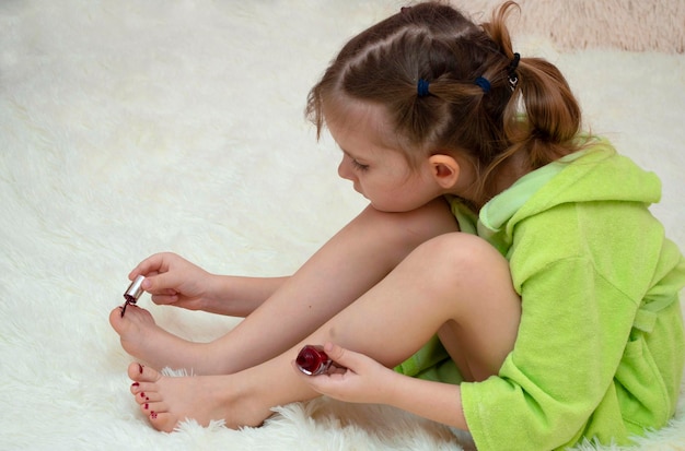 Photo little girl paints her toenails with bright red nail polish at home on a white blanket in bed.