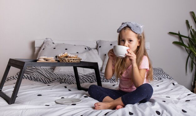 A little girl in pajamas is sitting on the bed with a tray of buns and a mug of tea