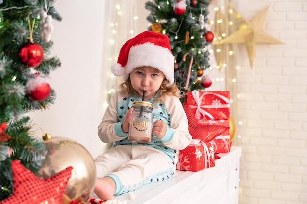 ココアミルクを飲むパジャマとサンタの帽子の少女！クリスマスの雰囲気に囲まれています。