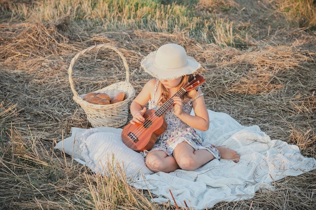 Little girl in panama hat play ukulele sitting on blanket on field among dry cut grass Outdoor walking and picnic