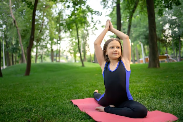 A little girl in the park sits on a mat and does yoga elements raising her hands forward