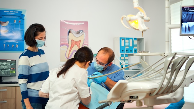 Little girl patient sitting in stomatological chair in dental office, visiting doctor for oral problems. Woman dentist with protective gloves and mask removes tooth decay drilling teeth during surgery