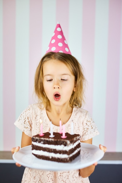 Bambina in tappo rosa che spegne le candeline su una torta al cioccolato di compleanno