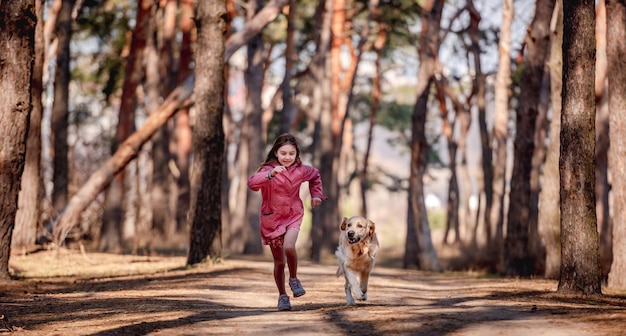 Little girl in pink coat smiling and running with golden retriever dog in the wood