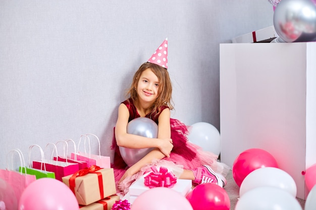 Little girl in pink dress and birthday hat sitting at the floor with many gift boxes and balloons