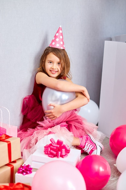 Little girl in pink dress and birthday hat sitting at the floor with many present gift box, balloons