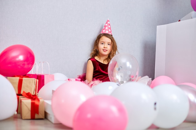 Bambina in abito rosa e cappello di compleanno seduta sul pavimento con molte scatole regalo presenti, palloncini