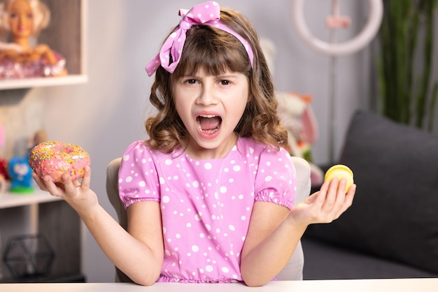 Little girl in pink dress holding donut and macaron in hands cute adolescent school girl plays