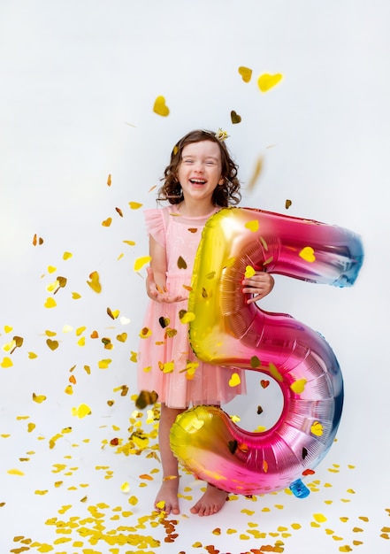 A little girl in a pink dress holds a multi-colored number five on a white background with gold confetti