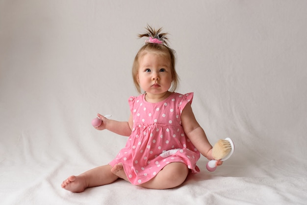 Маленькая девочка в розовом платье сидит с расческой в руке на белой поверхности