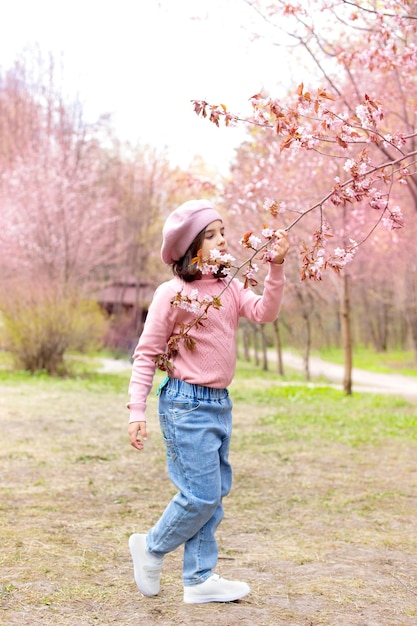 A little girl in a pink jumper and blue jeans sniffs a branch of cherry blossoms in the park during