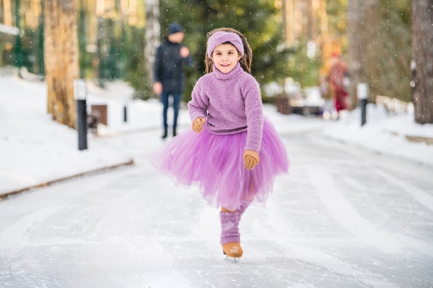 핑크 스웨터와 풀 스커트를 입은 어린 소녀는 화창한 겨울날 공원에 있는 야외 아이스링크에서 타기