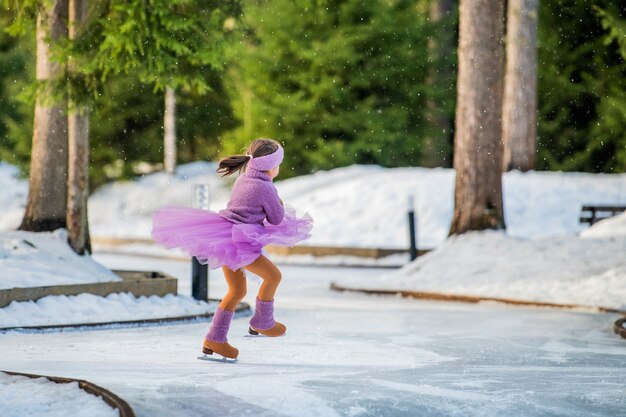 Little girl in pink sweater and full skirt rides on sunny winter day on an outdoor ice rink in park