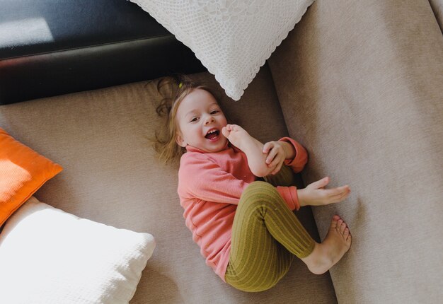 A little girl in a pink sweatshirt is lying on the sofa among the pillows with her legs up. Home games.