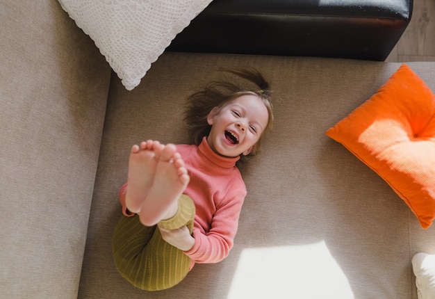 A little girl in a pink sweatshirt is lying on the sofa among the pillows with her legs up. Home games.