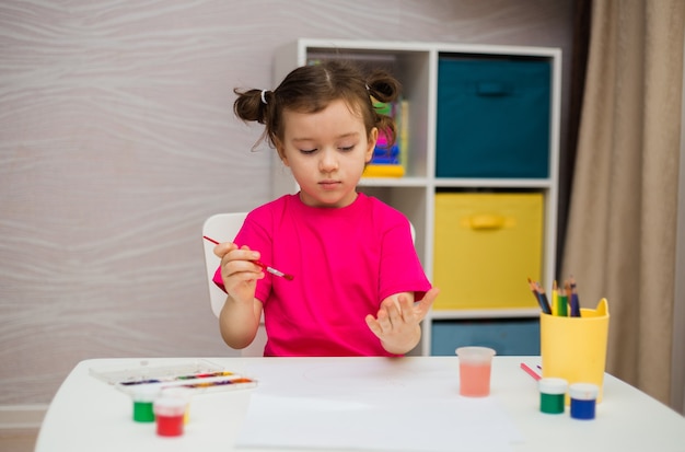 Little girl in a pink T-shirt sits at a table and draws with a brush and paints