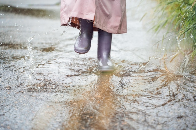 ピンクの防水レインコート紫のゴム長靴を着た少女は、雨の日の天候で通りの道路の水たまりを面白いジャンプします。春、秋、雨の後の子供の楽しみ、屋外レクリエーション活動