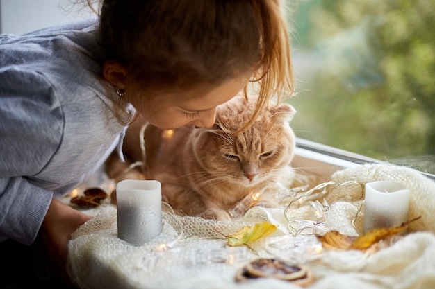 Little girl play with pet cat lying on window sill at home