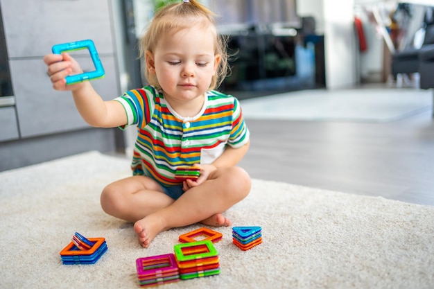 Маленькая девочка играет дома с красочными магнитными пластиковыми блоками, ребенок играет в развивающие игры и ...