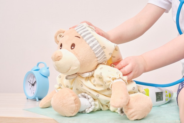 Little girl playing doctor and listening teddy bear with stethoscope alarm clock and pills on background