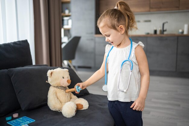 Little girl playing doctor with toys and teddy bear on the sofa in living room at home