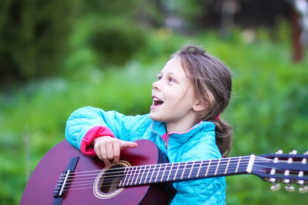 ギターを弾き、春の緑の牧草地で野外で歌う少女田舎の庭で幸せな子供