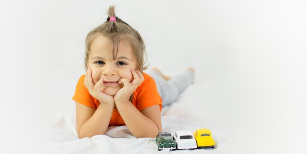 白いおもちゃの車で遊ぶ少女。白いシーツの上に置きます。笑顔。