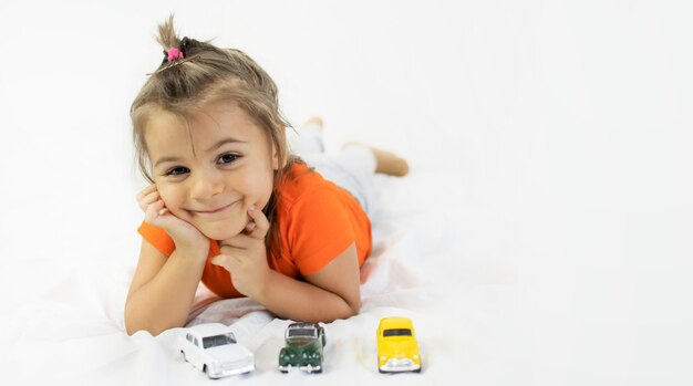 Little girl playing with white toy car. Laying on the white bedsheet. Smiling.