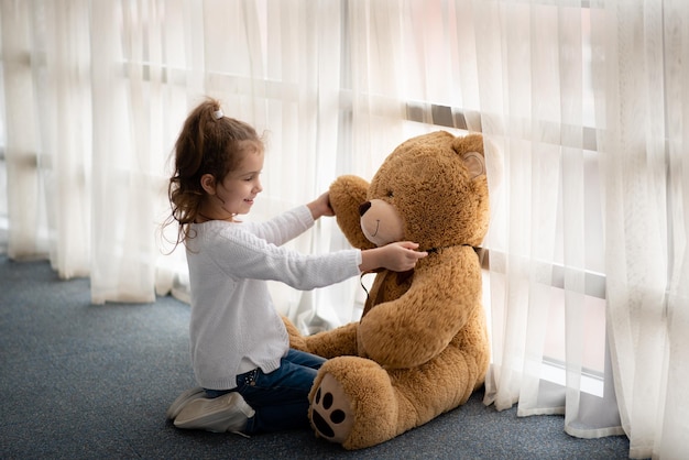 A little girl plays with a big teddy bear She is dressed in a white jumper Childhood Lifestile